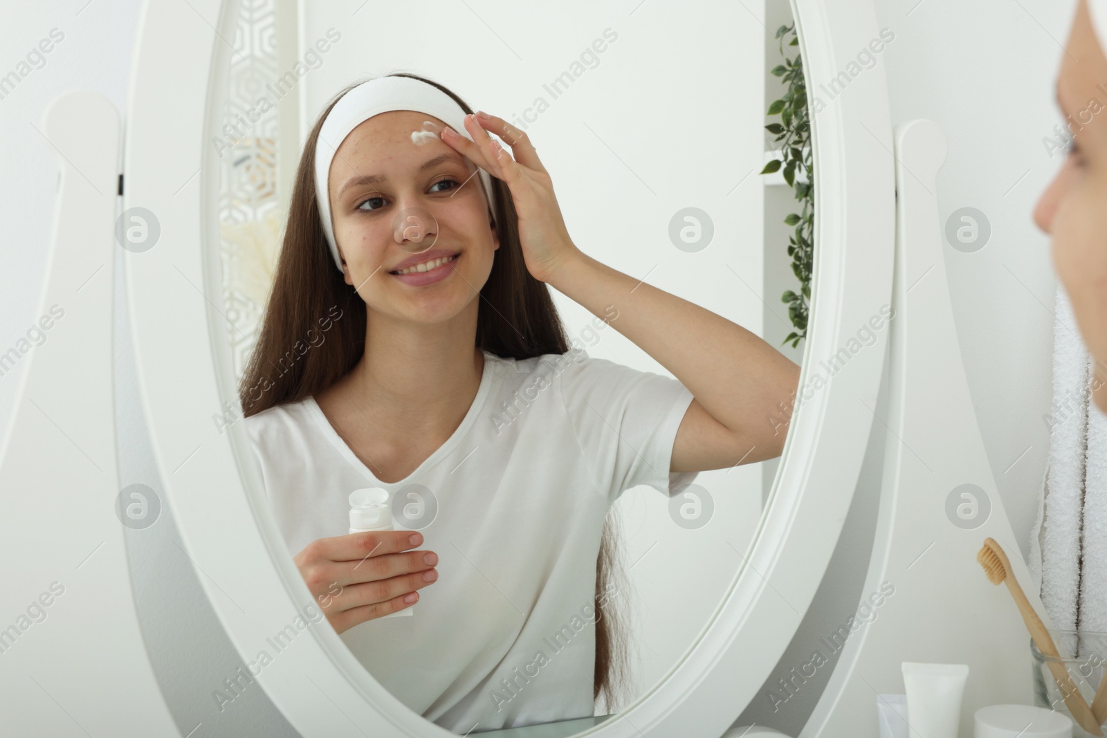 Photo of Teenage girl applying cream onto face near mirror indoors. Acne treatment