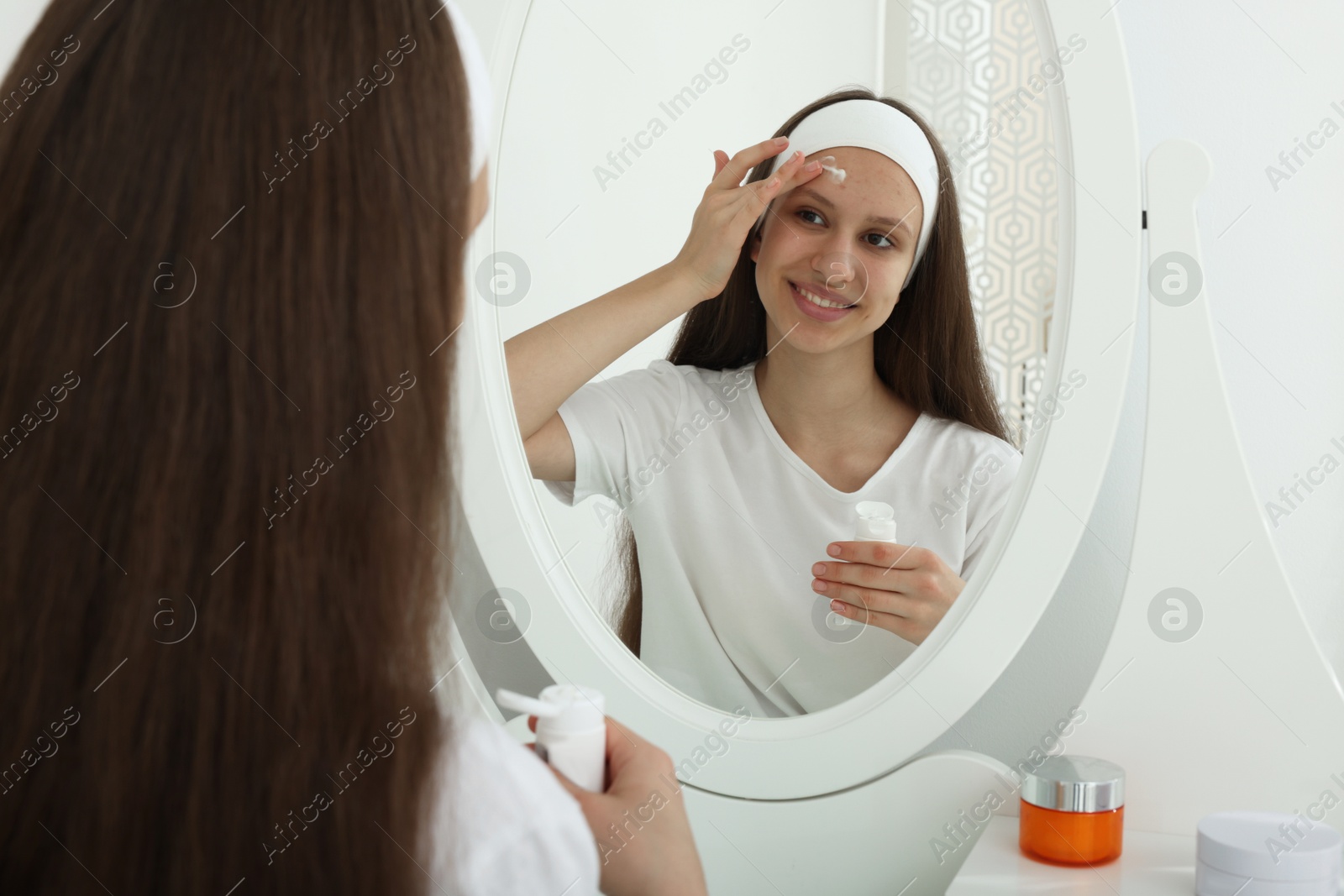 Photo of Teenage girl applying cream onto face near mirror indoors. Acne treatment