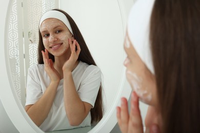 Photo of Teenage girl applying cream onto face near mirror indoors. Acne treatment