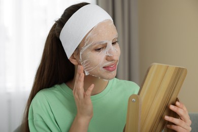 Photo of Teenage girl with sheet facial mask looking in mirror indoors. Acne treatment