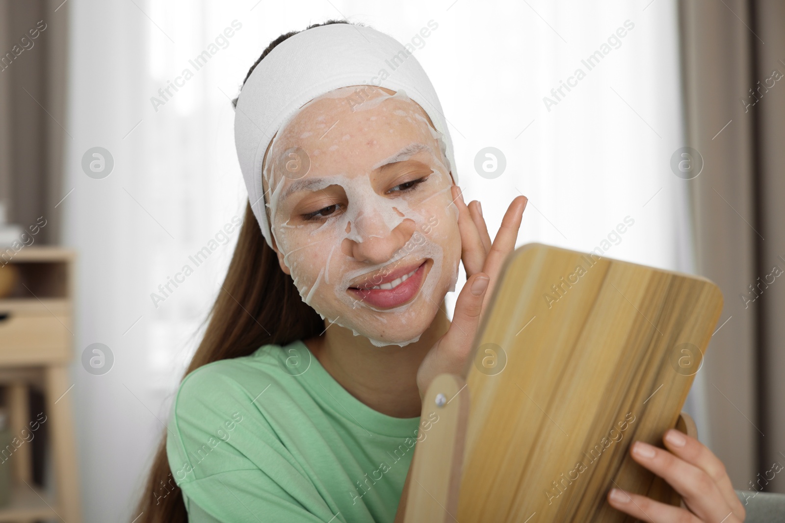 Photo of Teenage girl with sheet facial mask looking in mirror indoors. Acne treatment