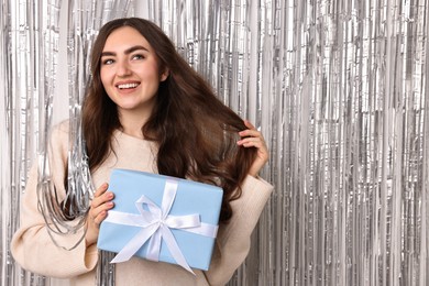 Photo of Happy young woman with gift box near silver foil curtain. Space for text