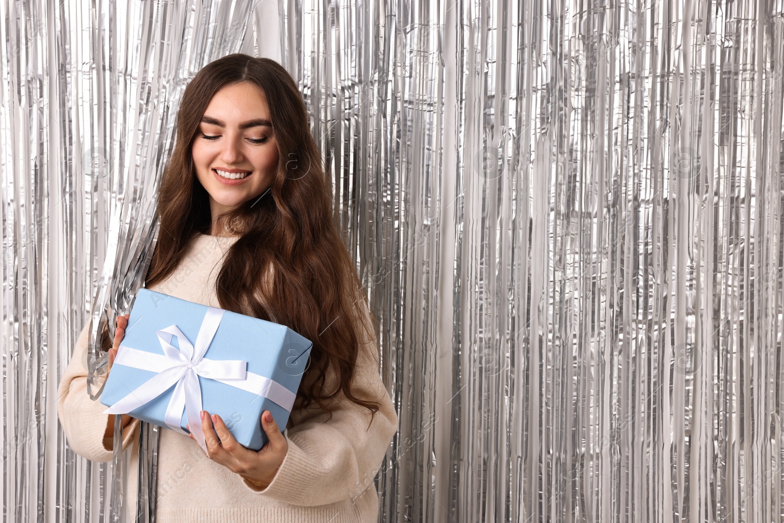 Photo of Happy young woman with gift box near silver foil curtain. Space for text