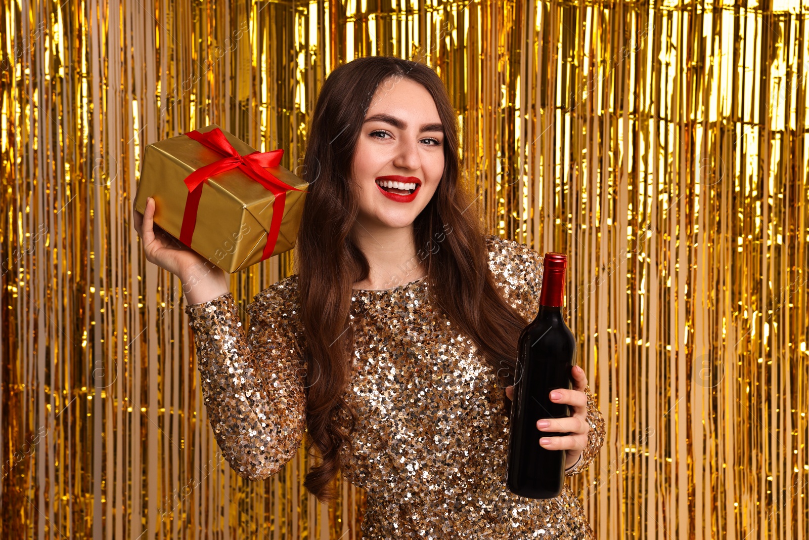 Photo of Happy young woman with gift box and bottle of wine against golden foil curtain