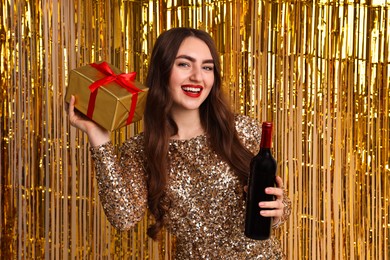 Photo of Happy young woman with gift box and bottle of wine against golden foil curtain