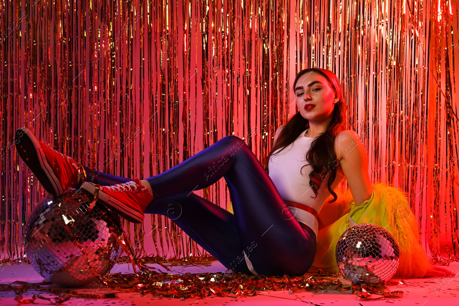 Photo of Beautiful young woman wearing retro outfit with tinsel and disco balls against golden foil curtain in neon lights