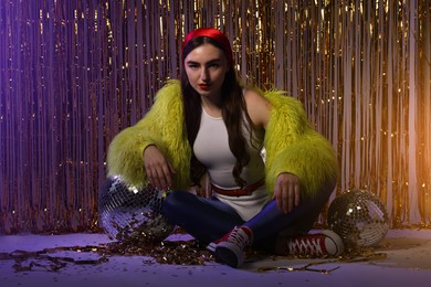 Photo of Beautiful young woman in retro outfit with shiny tinsel and disco balls indoors