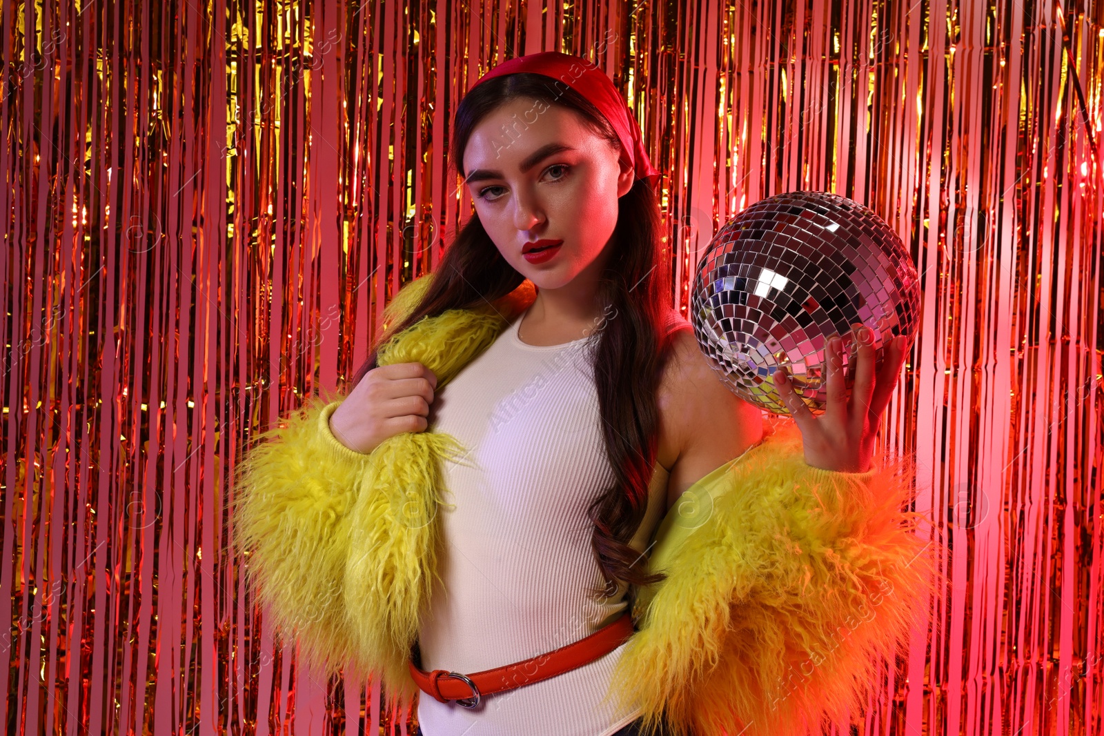 Photo of Beautiful young woman in faux fur coat with disco ball against golden foil curtain