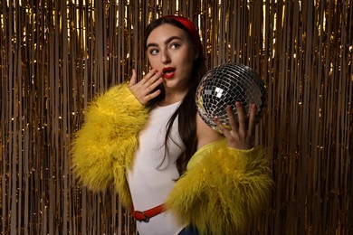 Photo of Beautiful young woman in faux fur coat with disco ball against golden foil curtain