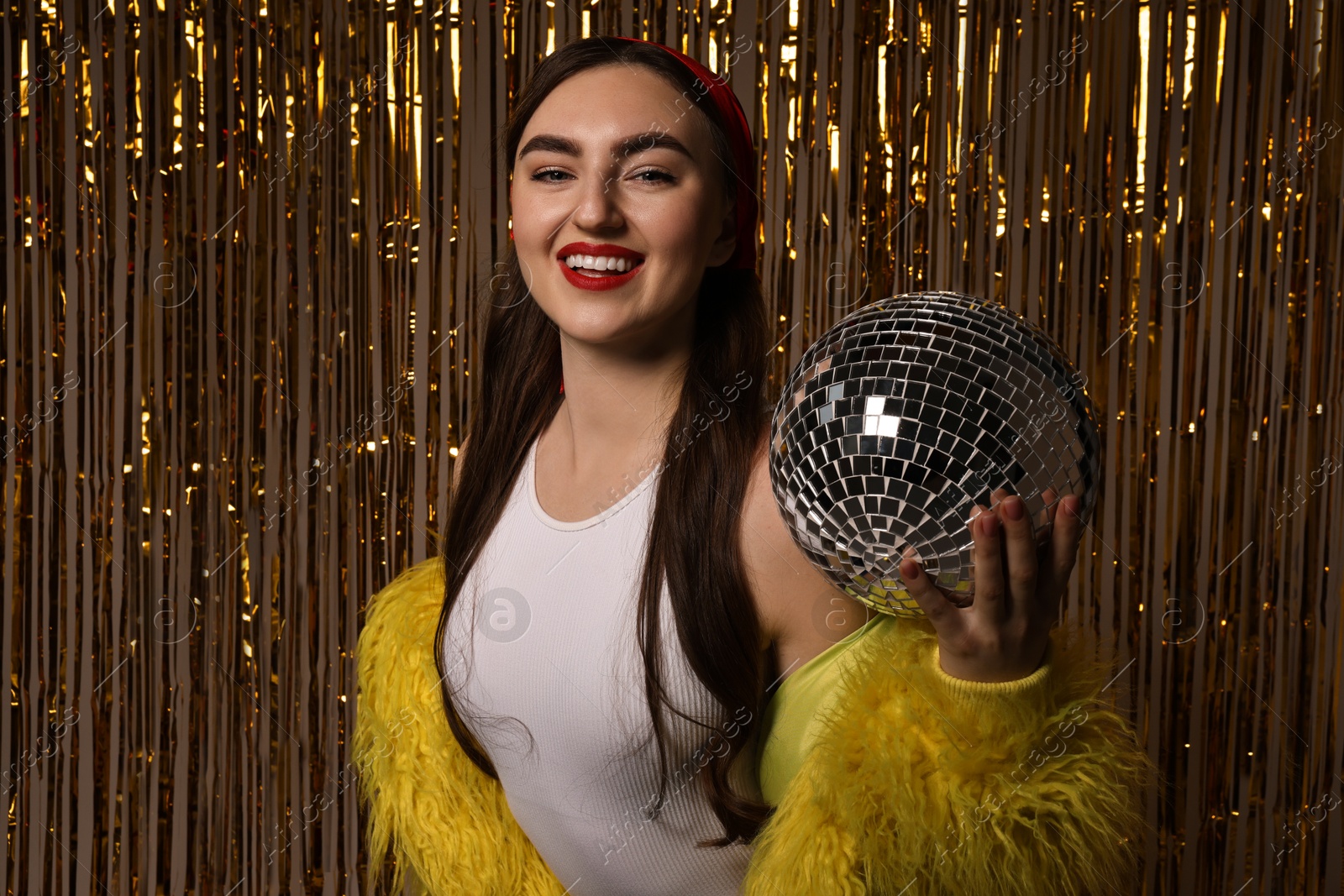 Photo of Beautiful young woman in faux fur coat with disco ball against golden foil curtain