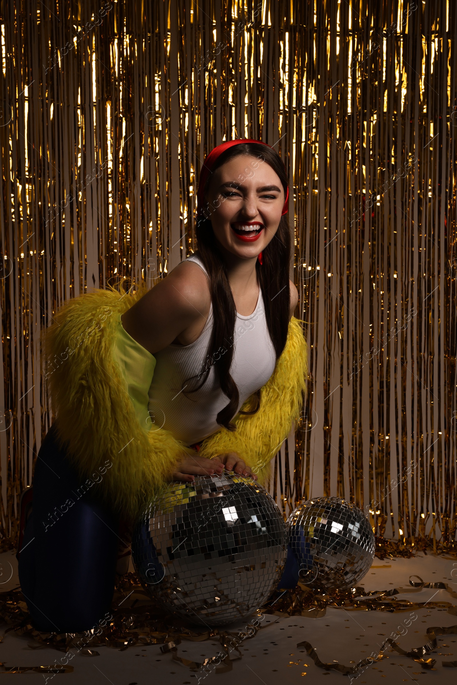 Photo of Beautiful young woman in retro outfit with tinsel and disco balls against golden foil curtain indoors