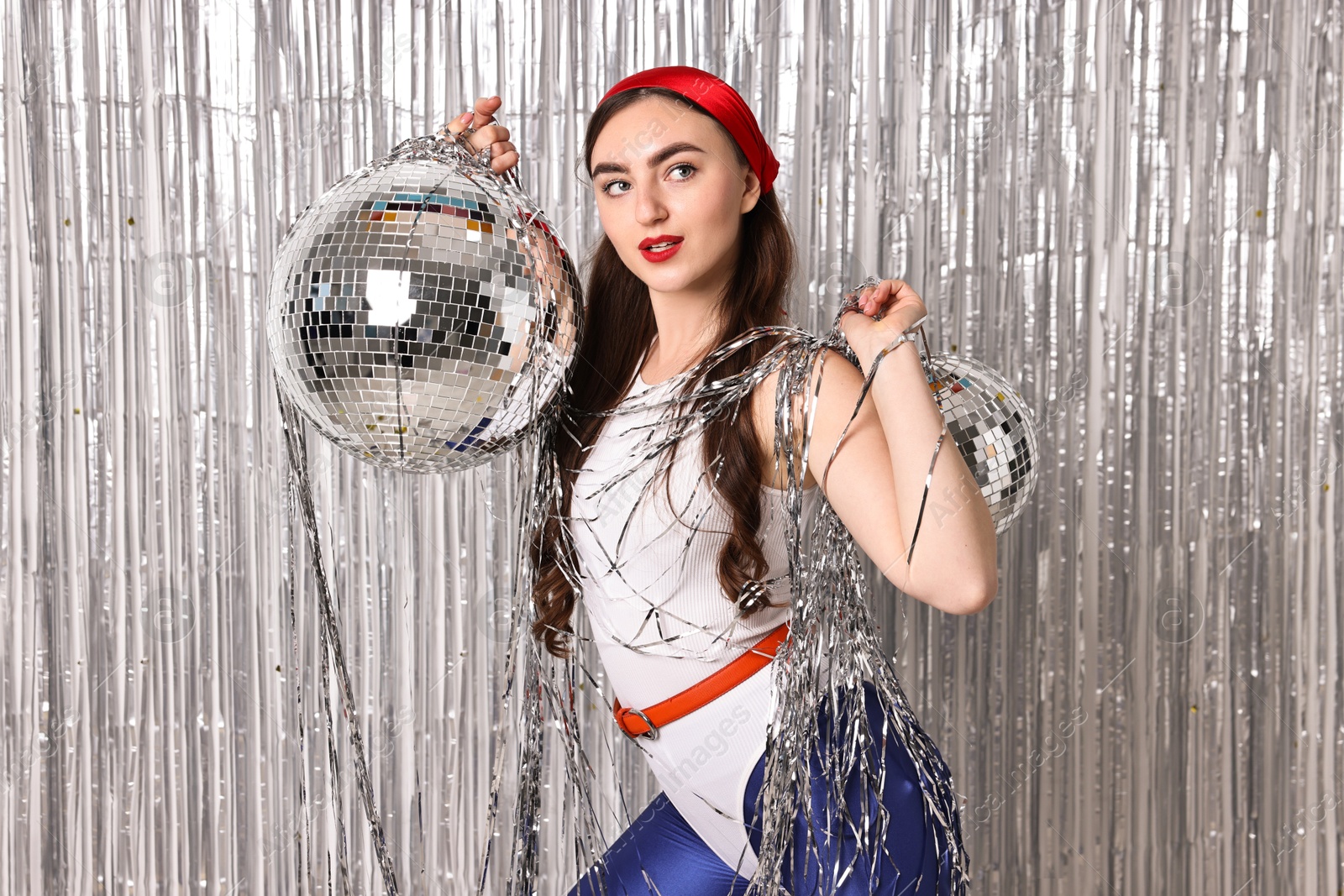 Photo of Beautiful young woman in retro outfit with disco balls against silver foil curtain