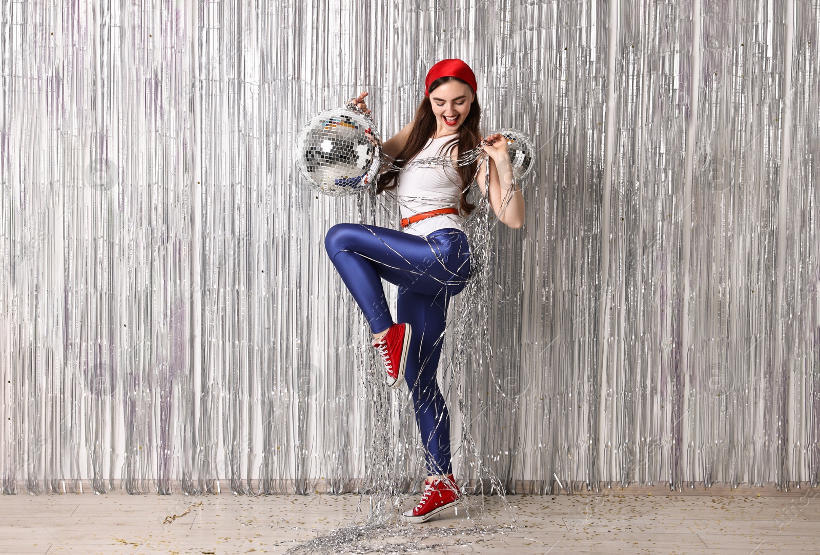 Photo of Beautiful young woman in retro outfit with disco balls against silver foil curtain indoors