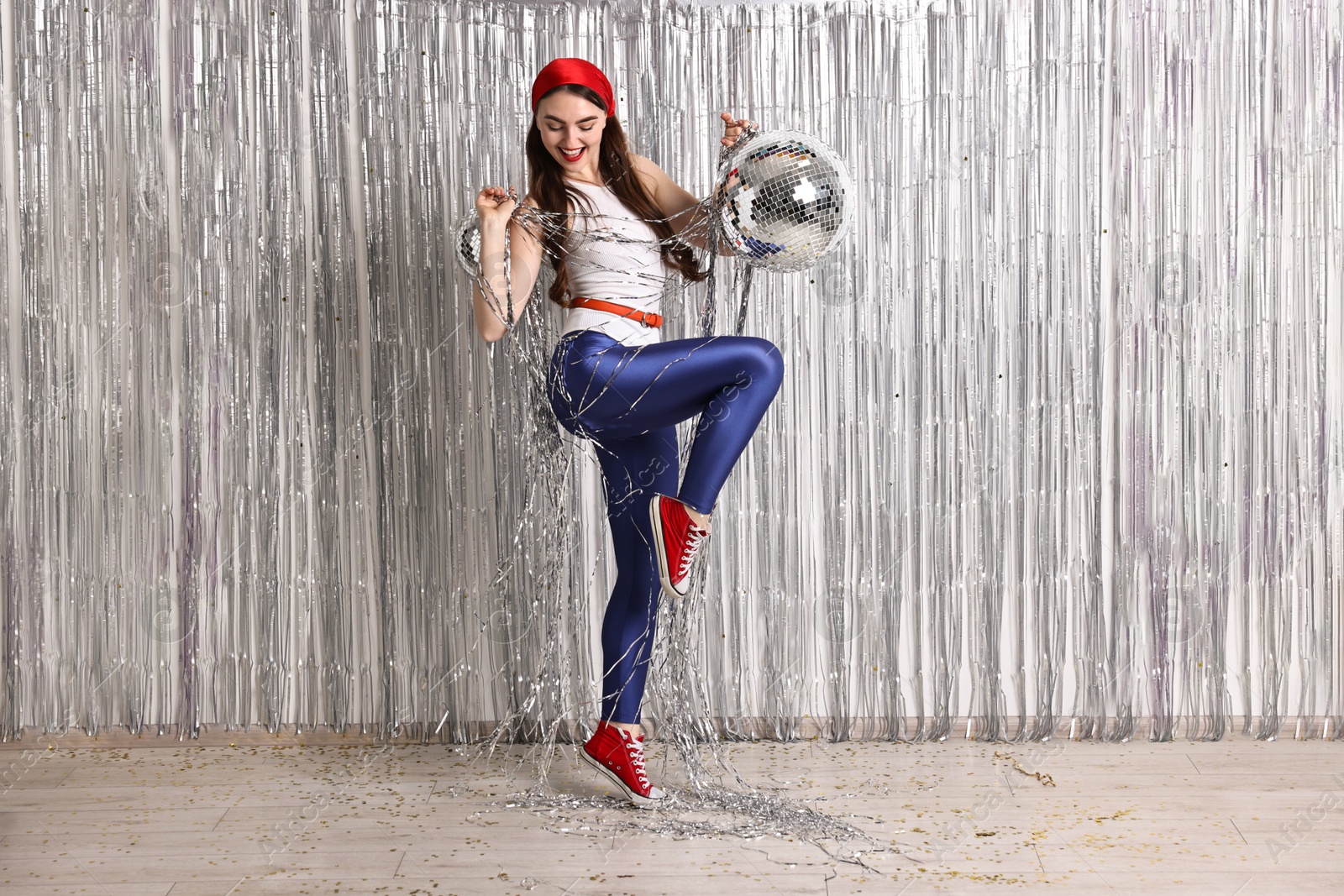 Photo of Beautiful young woman in retro outfit with disco balls against silver foil curtain indoors