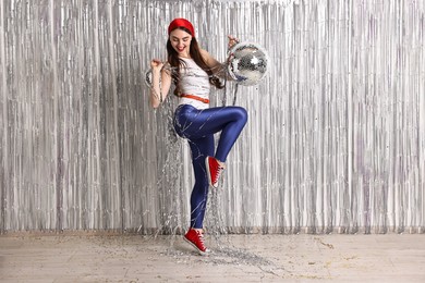 Photo of Beautiful young woman in retro outfit with disco balls against silver foil curtain indoors