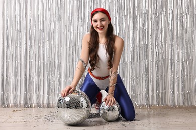 Photo of Beautiful young woman in retro outfit with disco balls against silver foil curtain indoors