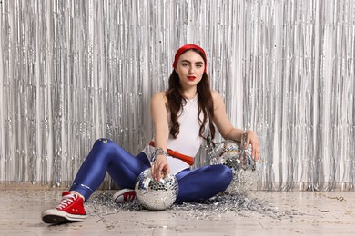 Photo of Beautiful young woman in retro outfit with disco balls against silver foil curtain indoors