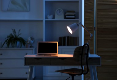 Photo of Comfortable workplace with laptop and lamp on table indoors