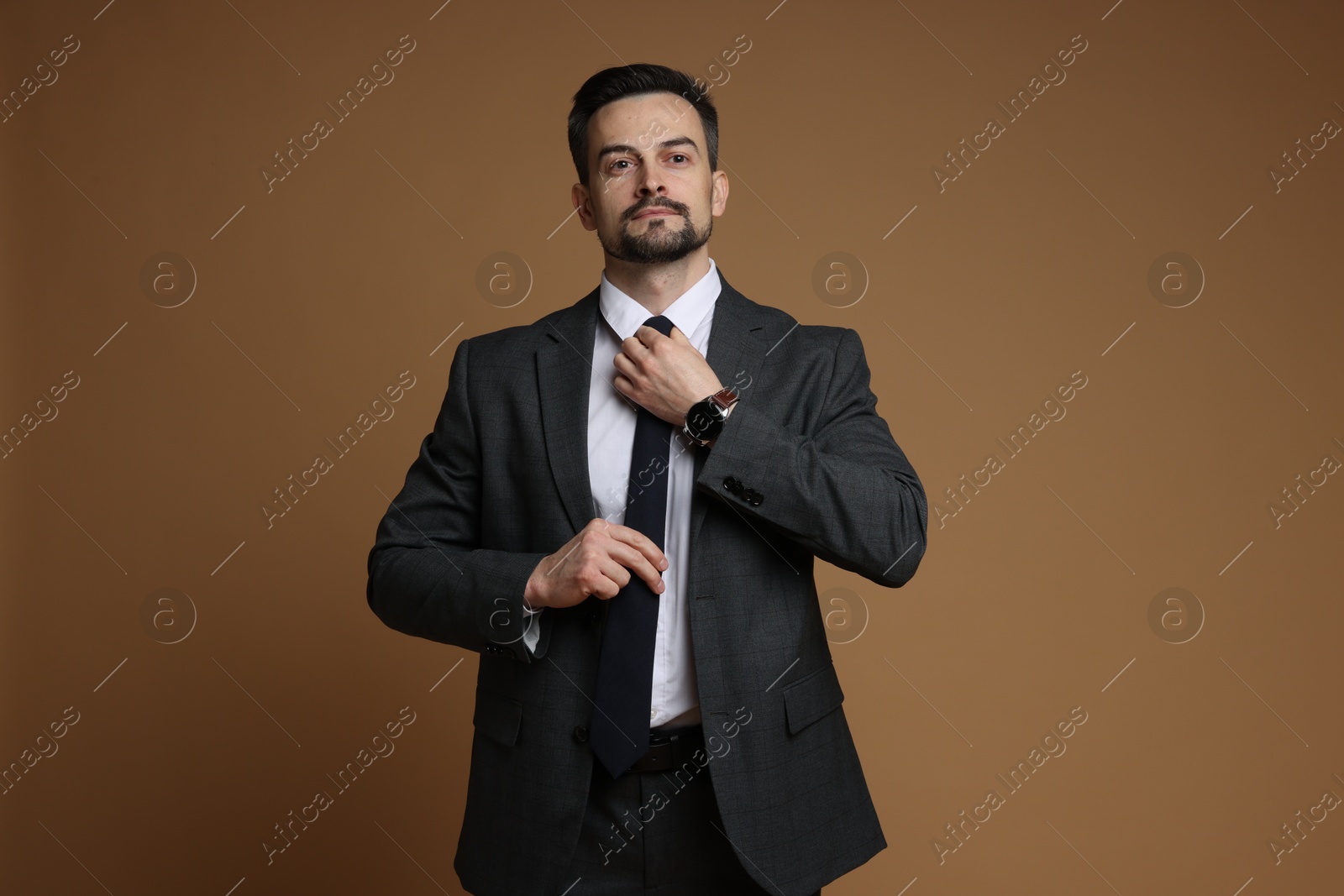 Photo of Confident man in classic suit straightening tie on brown background