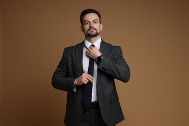 Photo of Confident man in classic suit straightening tie on brown background