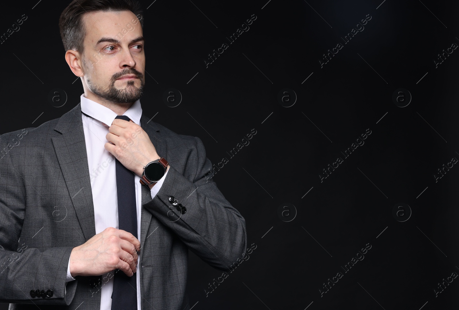 Photo of Confident man in classic suit straightening tie on black background. Space for text