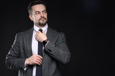 Photo of Confident man in classic suit straightening tie on black background. Space for text