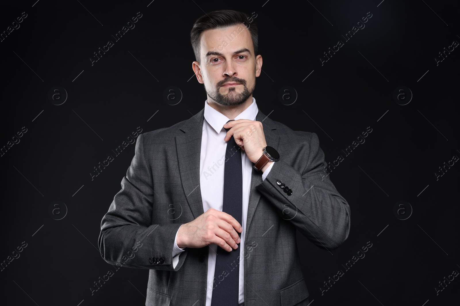 Photo of Confident man in classic suit straightening tie on black background