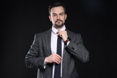 Photo of Confident man in classic suit straightening tie on black background