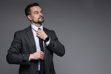 Photo of Confident man in classic suit straightening tie on grey background. Space for text