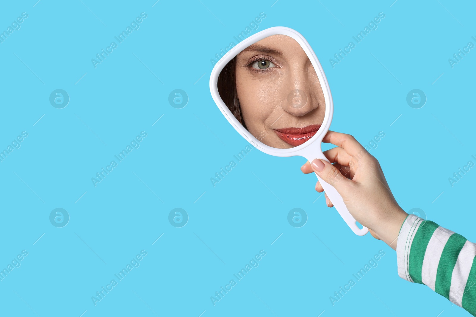 Image of Beautiful woman looking in handheld mirror on light blue background, closeup