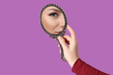 Image of Beautiful woman looking in handheld mirror on violet background, closeup