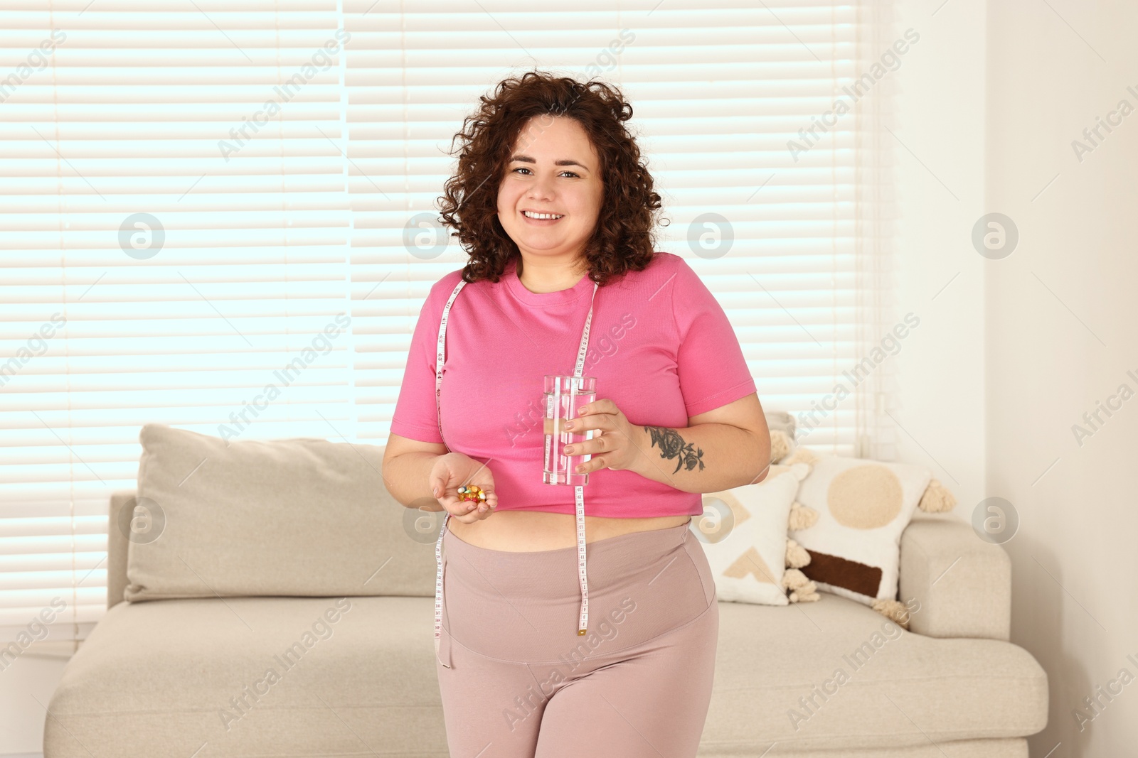 Photo of Happy plus size woman holding pile of weight loss supplements and glass with water at home