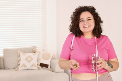 Photo of Happy plus size woman holding pile of weight loss supplements and glass with water at home. Space for text