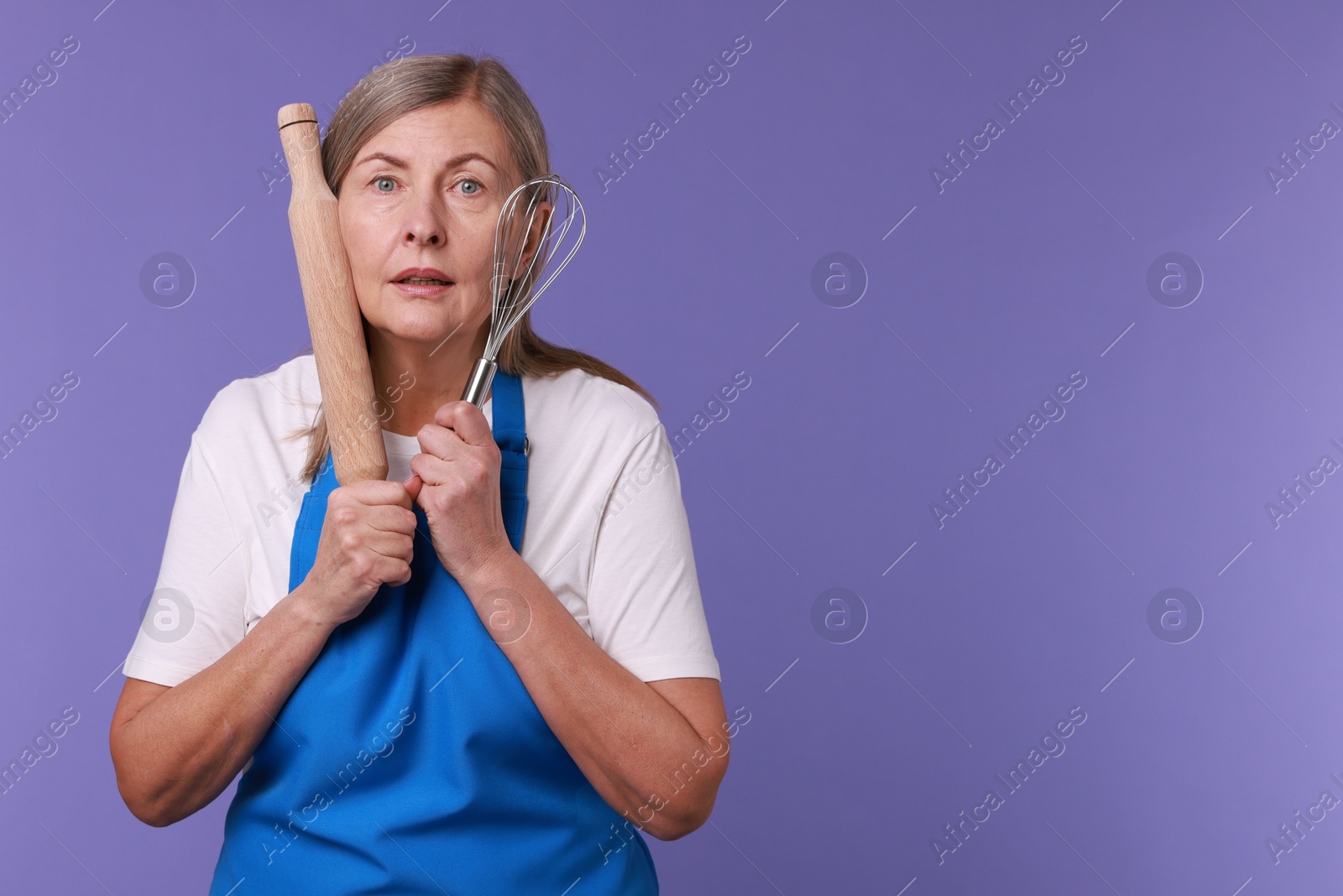 Photo of Surprised woman with rolling pin and whisk on violet background. Space for text