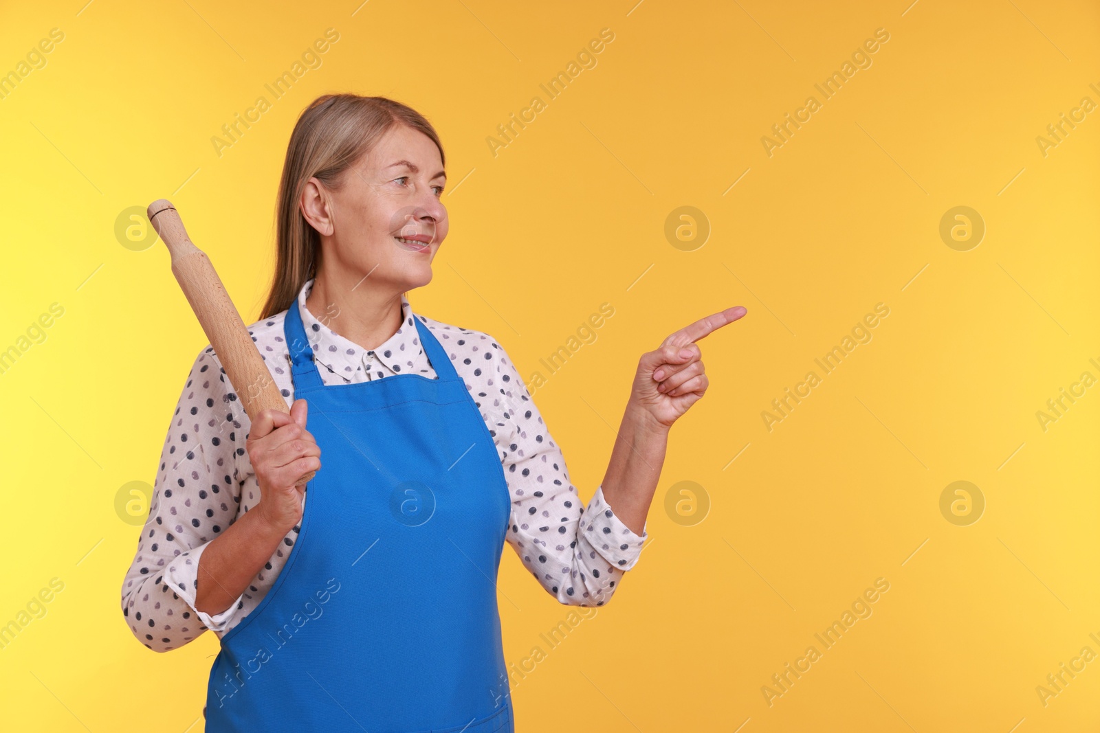 Photo of Happy woman with rolling pin pointing at something on yellow background. Space for text