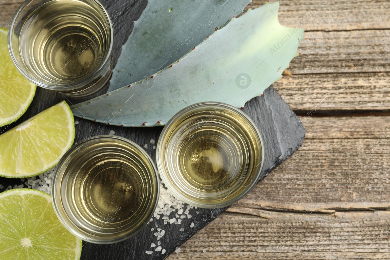 Photo of Mexican tequila shots, slices of lime, salt and agave leaves on wooden table, flat lay. Space for text