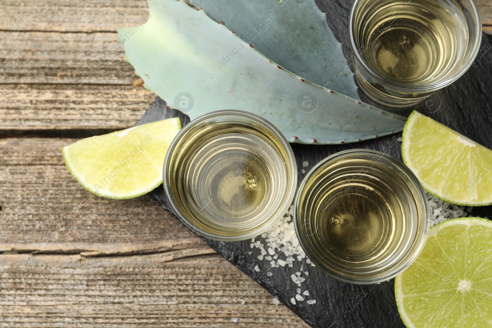 Photo of Mexican tequila shots, slices of lime, salt and agave leaves on wooden table, flat lay. Space for text