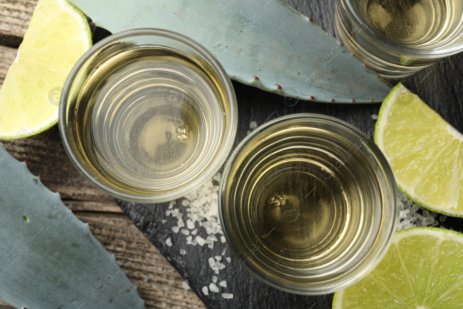 Photo of Mexican tequila shots, slices of lime, salt and agave leaves on wooden table, flat lay