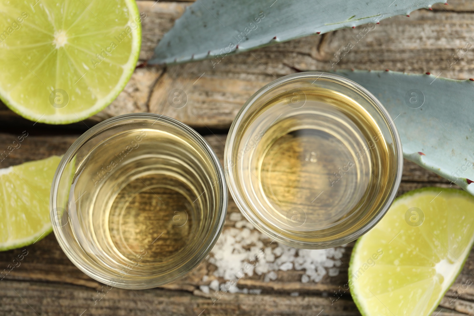 Photo of Mexican tequila shots, slices of lime, salt and agave leaves on wooden table, flat lay