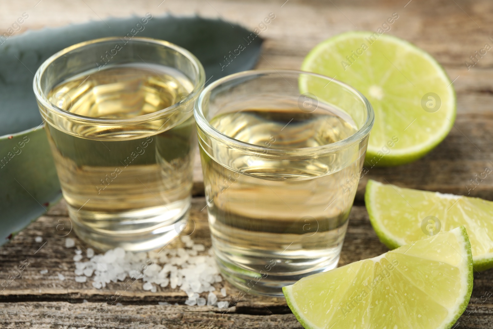 Photo of Mexican tequila shots, slices of lime, salt and agave leaves on wooden table, closeup