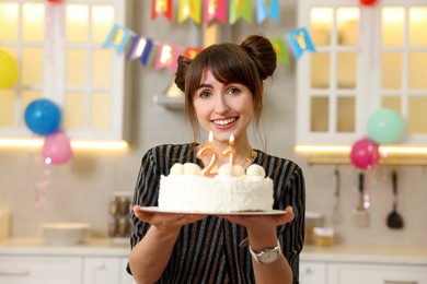 Photo of Coming of age party - 21st birthday. Happy young woman holding tasty cake with number shaped candles at home