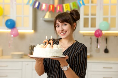 Coming of age party - 21st birthday. Happy young woman holding tasty cake with number shaped candles at home