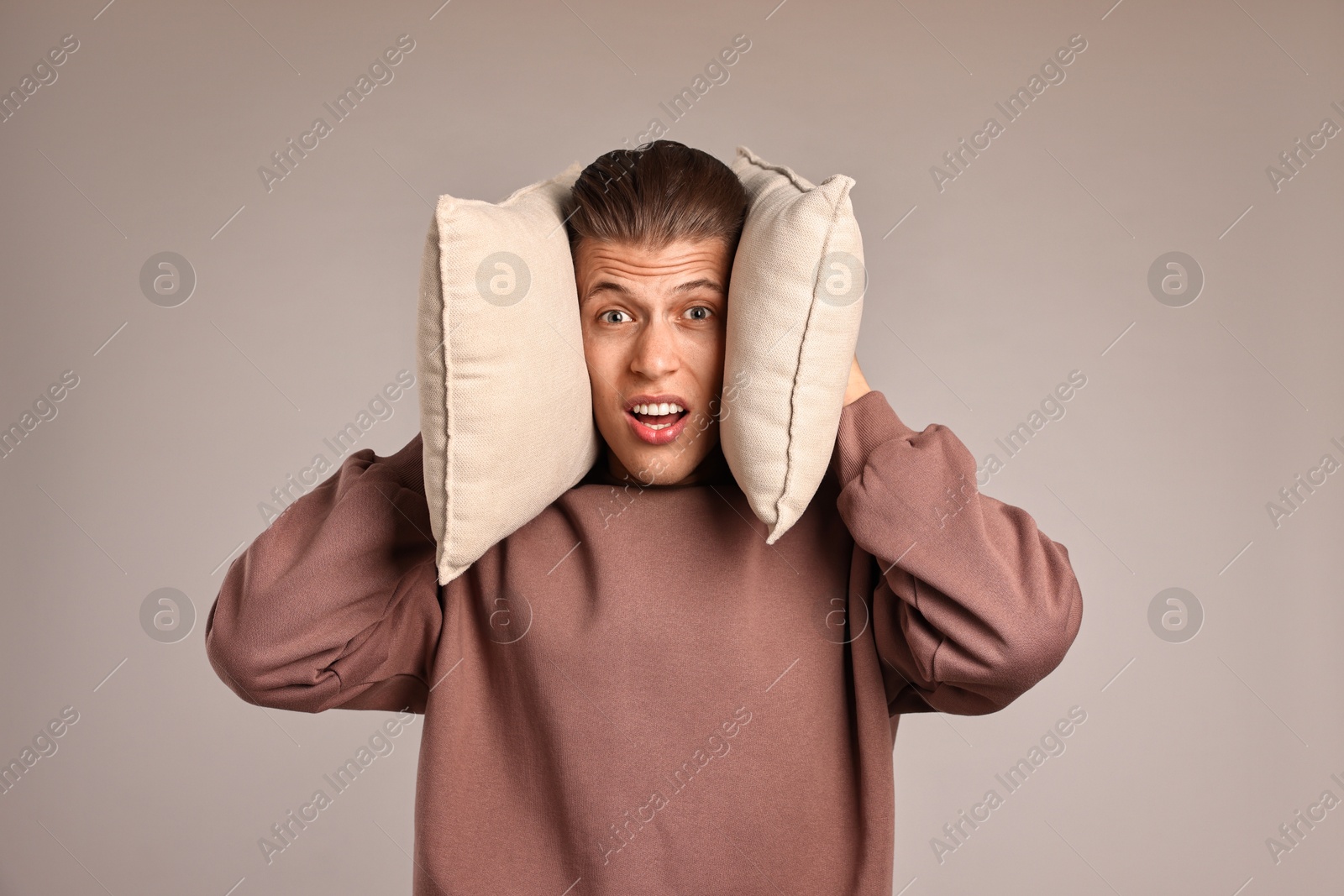 Photo of Annoyed young man covering his ears with pillows due to loud sound on light grey background