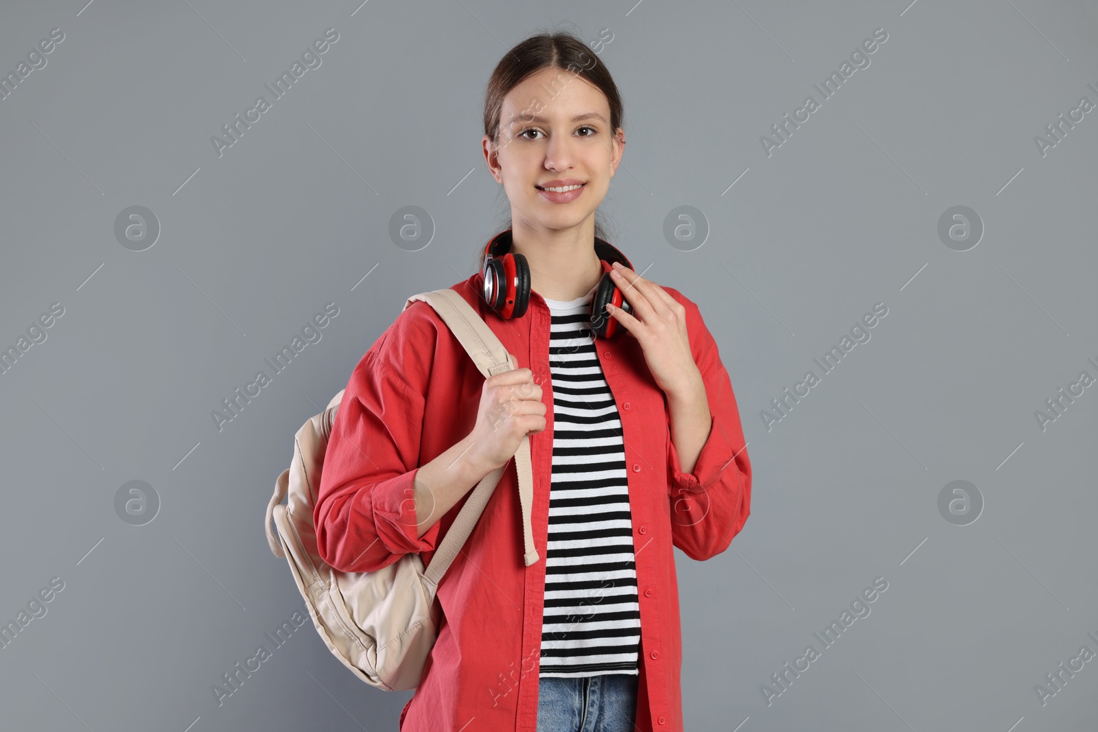 Photo of Portrait of smiling teenage girl with backpack and headphones on grey background