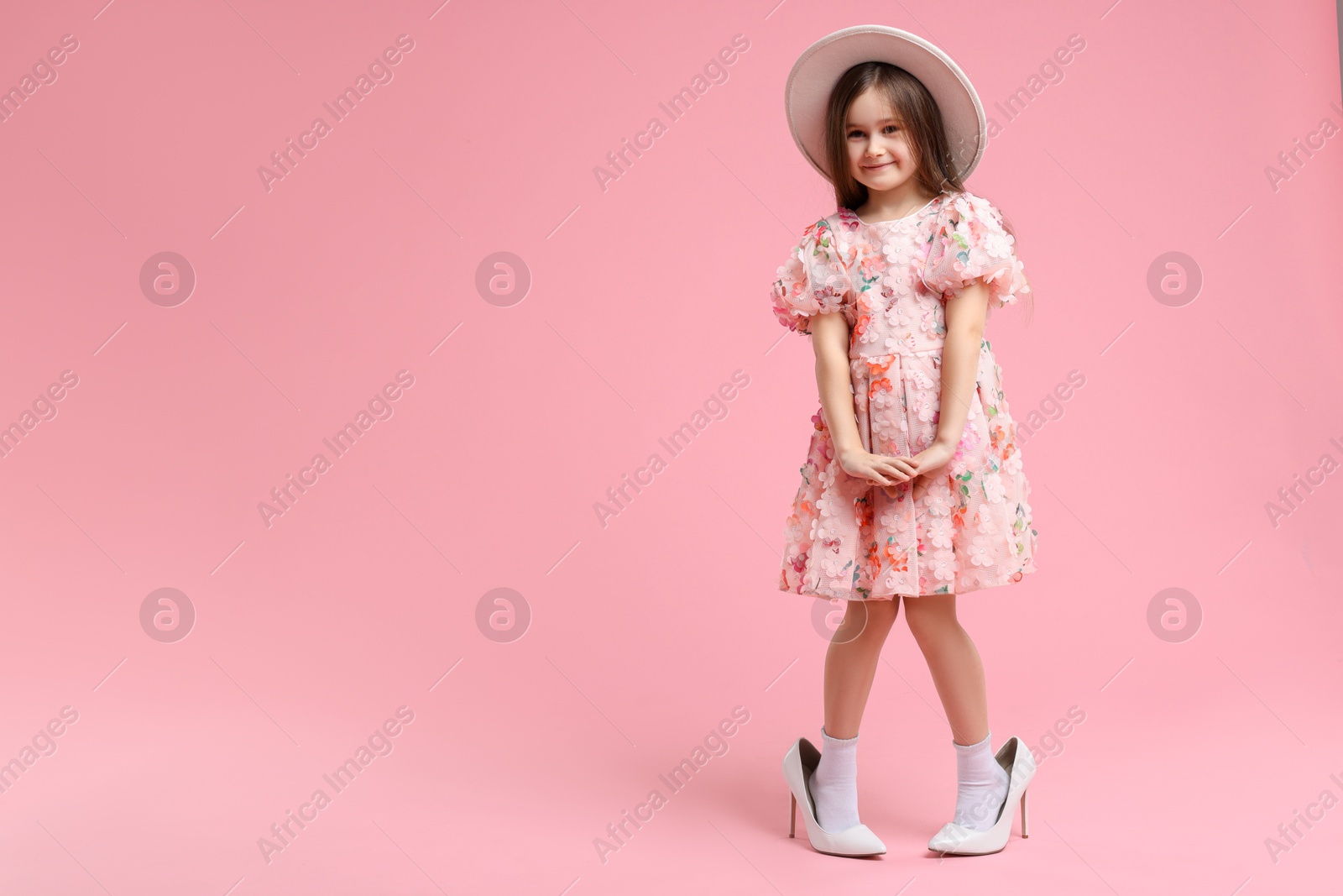 Photo of Stylish little girl wearing oversized high heeled shoes on pink background, space for text