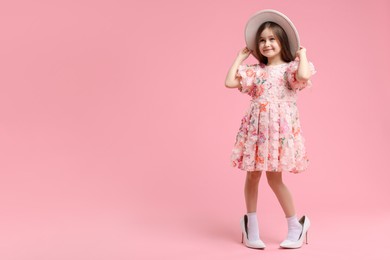 Photo of Stylish little girl wearing oversized high heeled shoes on pink background, space for text