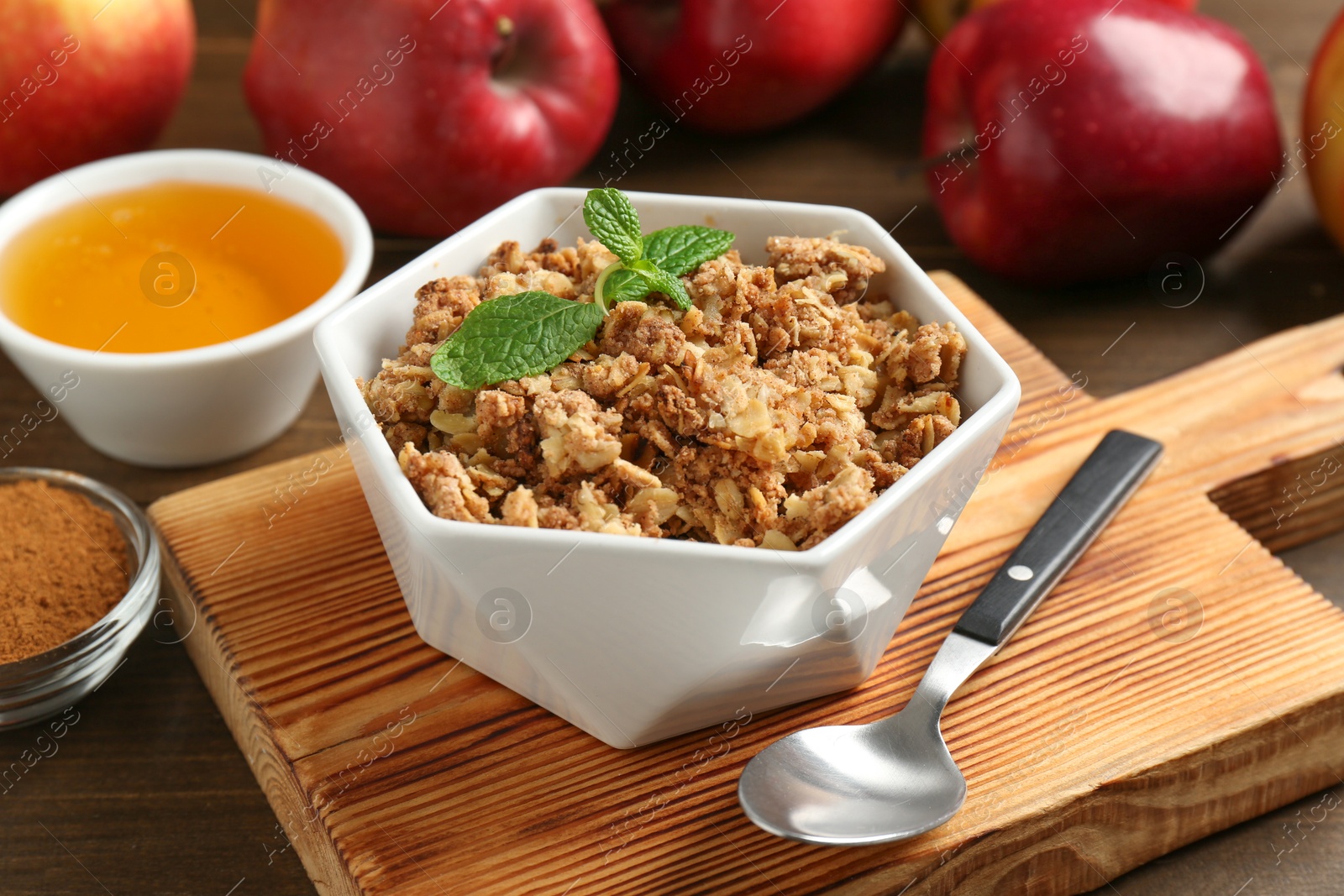 Photo of Tasty apple crisp in bowl, ingredients and spoon on wooden table, closeup