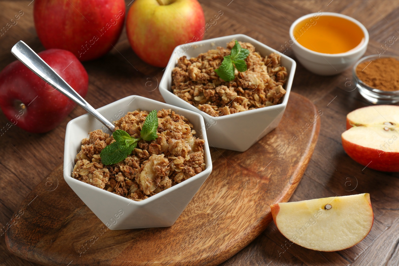 Photo of Tasty apple crisp in bowls, ingredients and spoon on wooden table, closeup