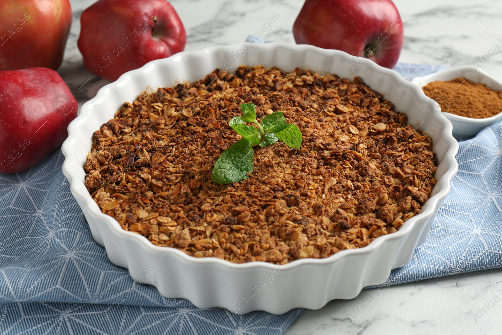 Photo of Tasty apple crisp in baking dish, fresh fruits and cinnamon on white marble table, closeup