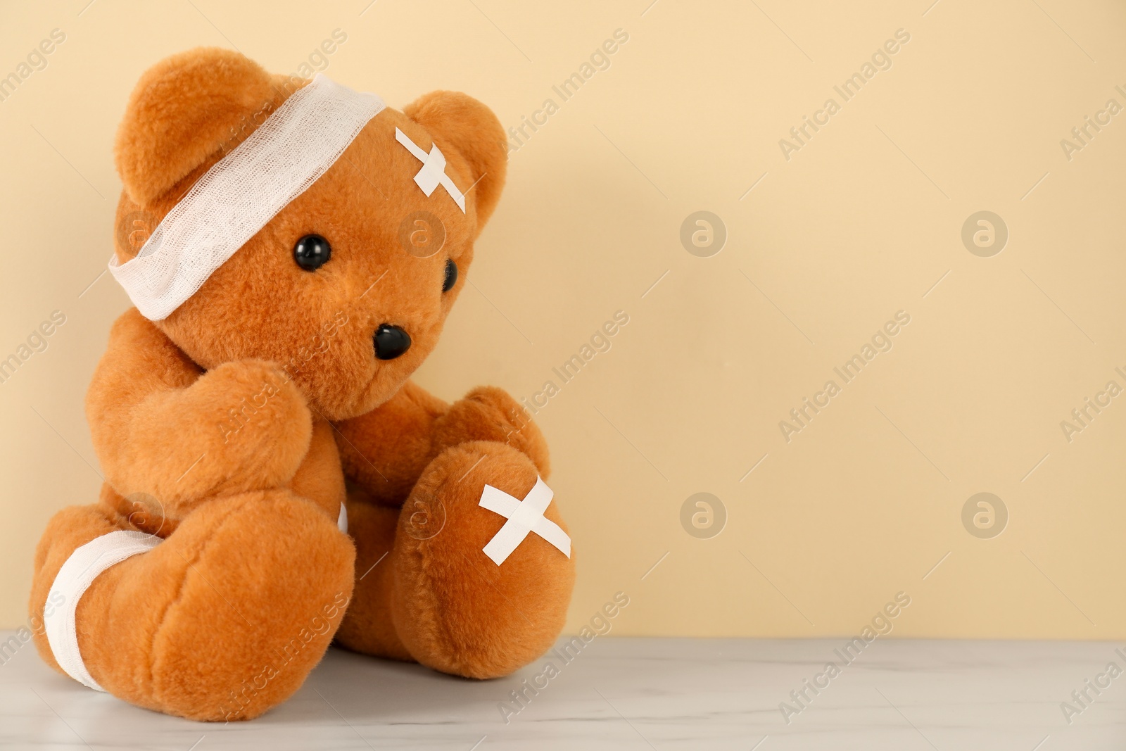 Photo of Teddy bear with bandages and sticking plasters on white marble table against beige background, space for text